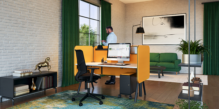 In einem gemütlichen Homeoffice mit einer markanten Backsteinwand und einem dunklen Holzboden führt ein Mann gerade ein Telefongespräch. In der Mitte des Raums steht ein stilvoller Schreibtisch mit einer orangefarbenen Akustikabschirmung. Daneben befindet sich ein HUGY-Bürostuhl mit einem eleganten schwarzen Gestell.